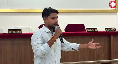 Atul Kumar Verma addressing students with a microphone, stage placards behind him.